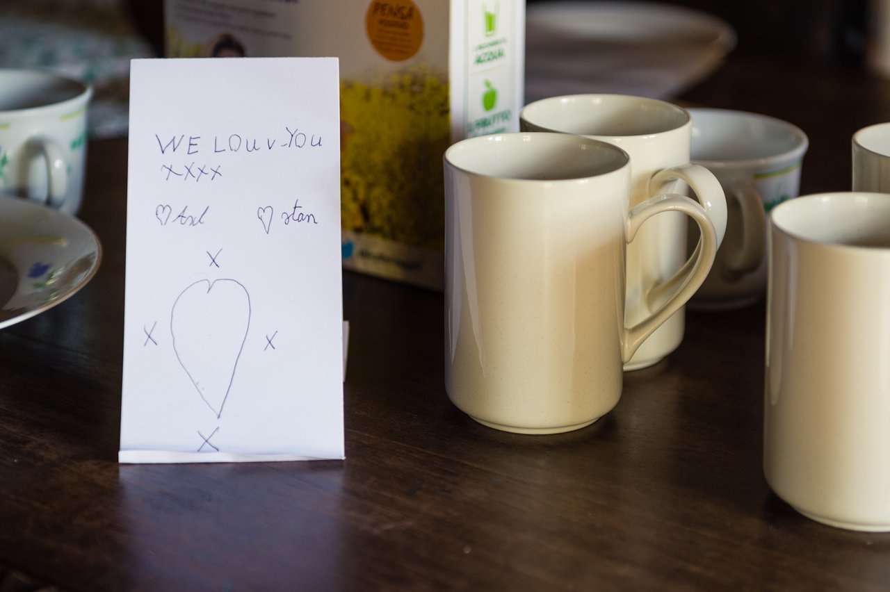 A handwritten note with a heart and message of love stands on a table surrounded by coffee mugs.
