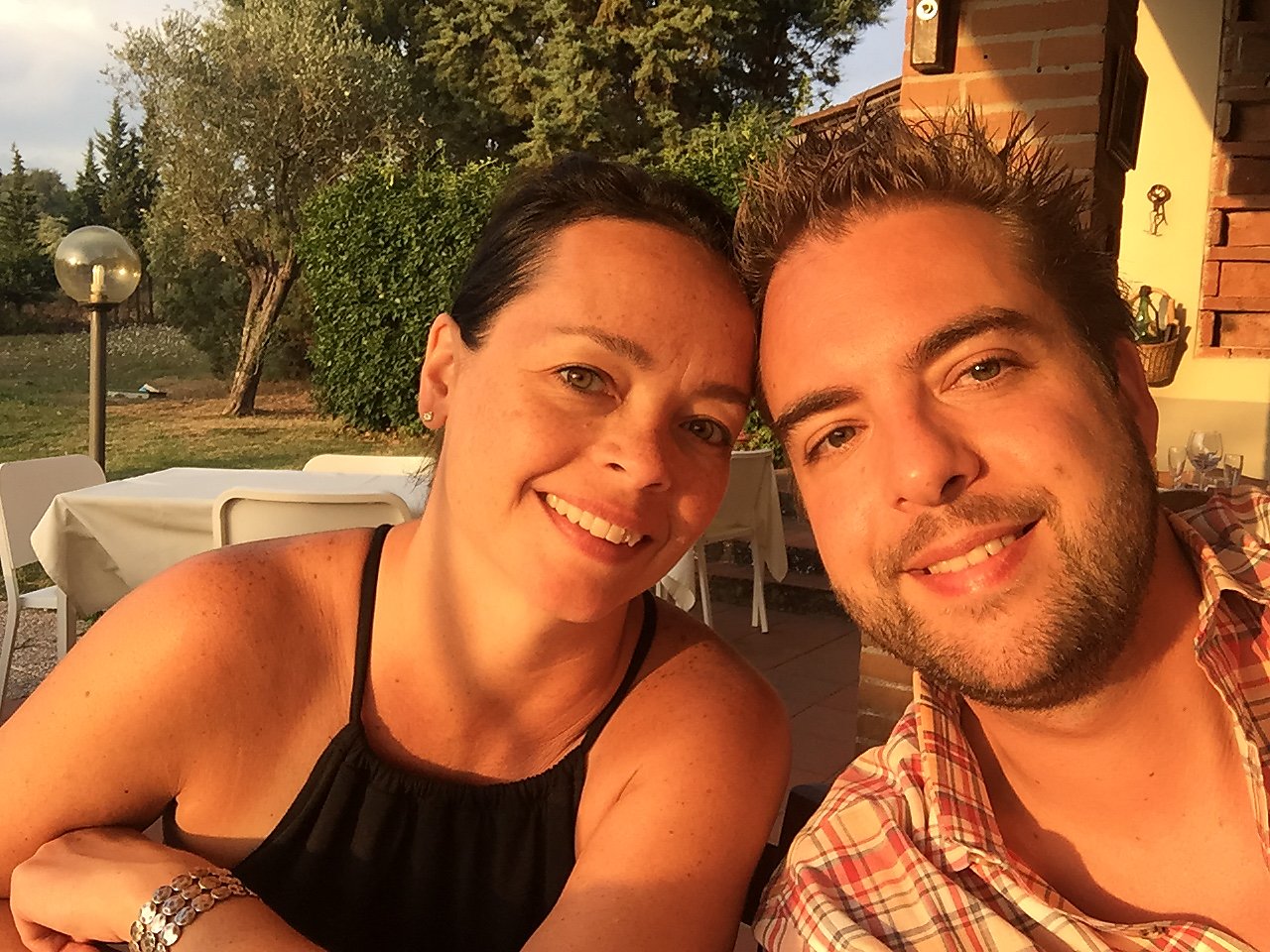 A smiling couple takes a close-up selfie at an outdoor restaurant during sunset.