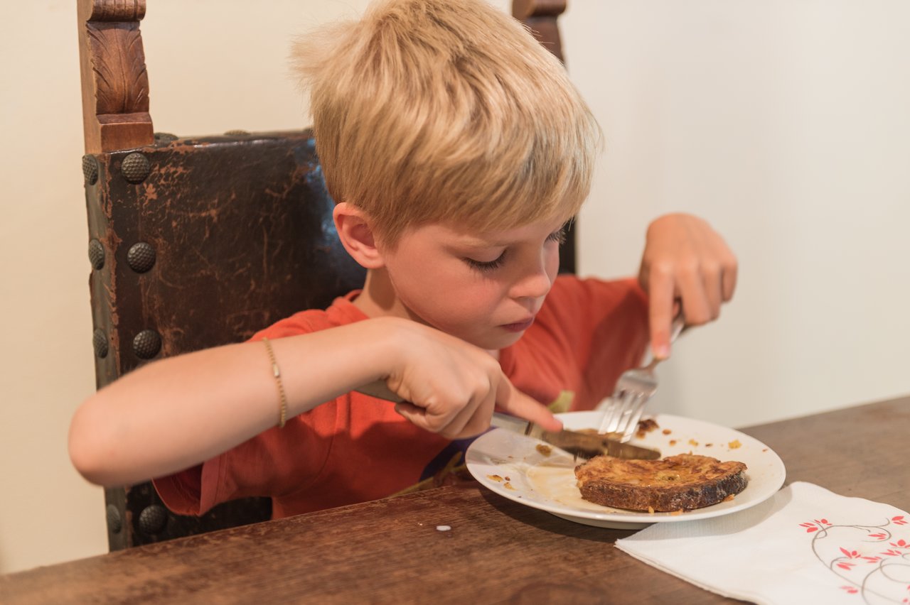 A child in an orange shirt cuts into a piece of French toast on a white plate at a table.