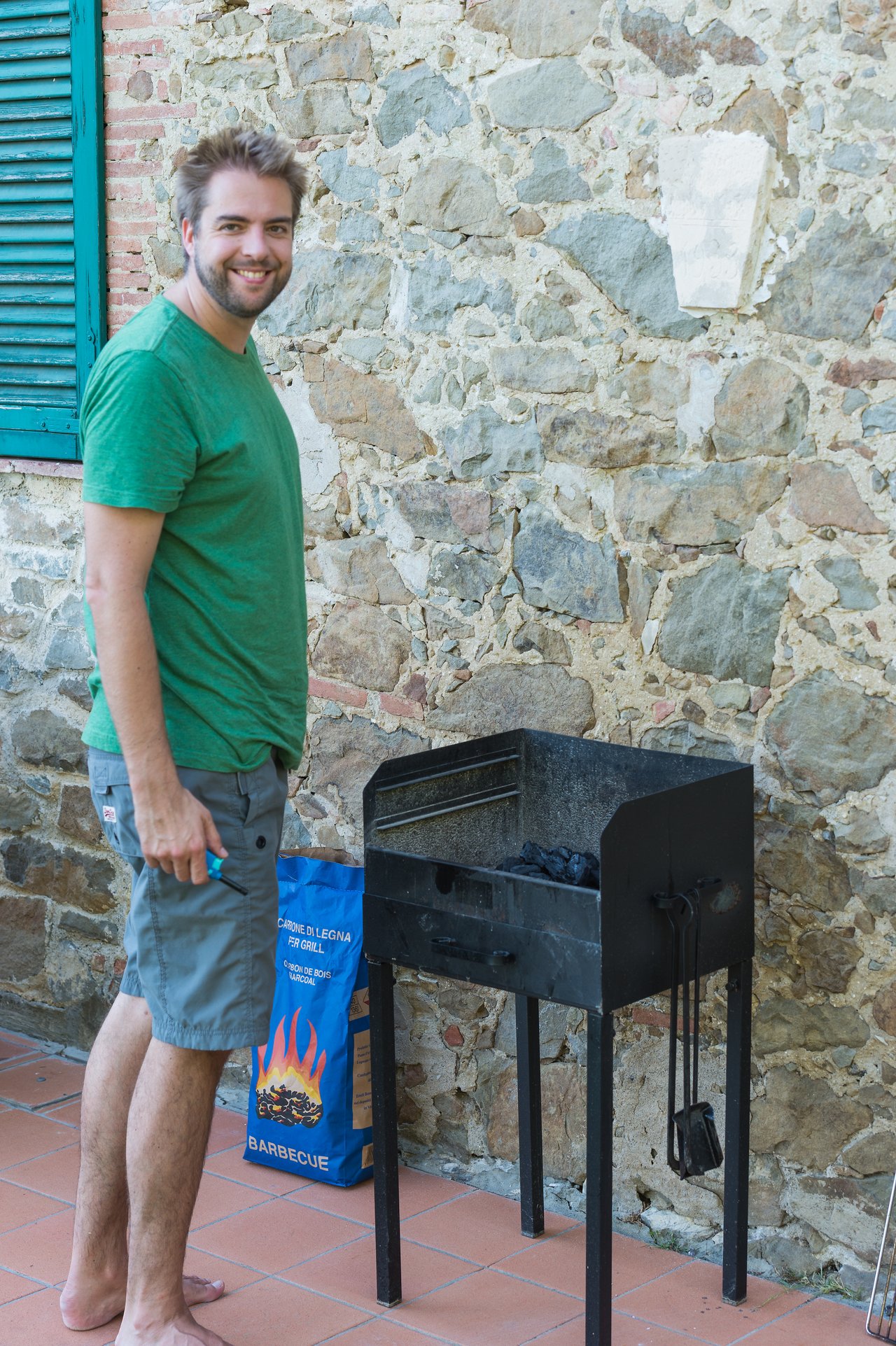 A man in a green shirt stands next to a charcoal grill, holding a lighter and preparing to start a fire.