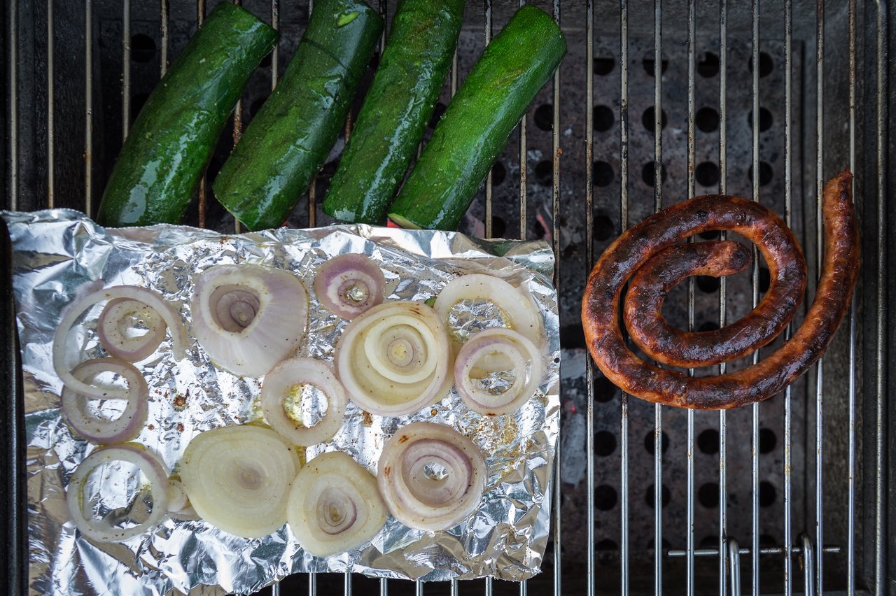 Sliced onions on foil, zucchini, and a coiled sausage cooking on a grill.