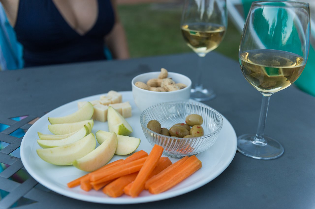 A plate with apple slices, carrot sticks, cheese, olives, and crackers, alongside two glasses of white wine.
