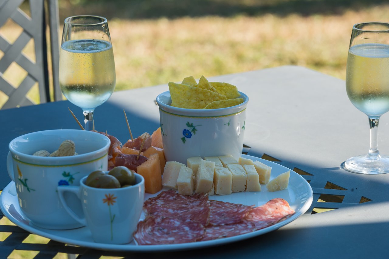 A plate with cheese, salami, olives, and snacks on an outdoor table with two glasses of white wine.