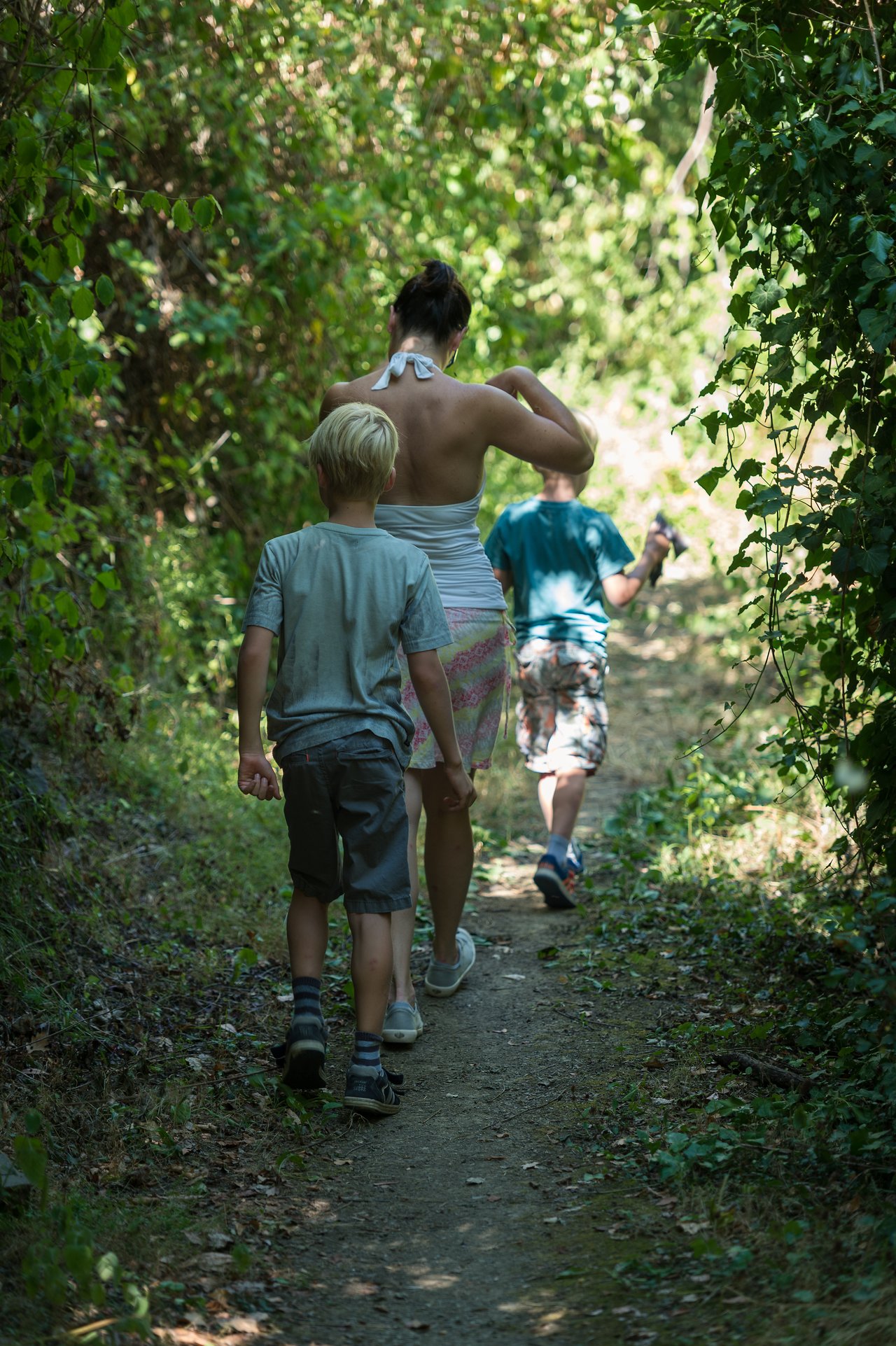 A woman and two children walk up a narrow dirt path surrounded by green foliage.