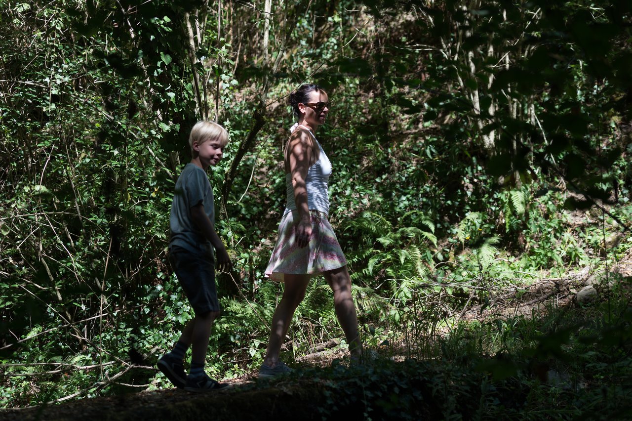 A woman and a child walk uphill on a forest trail, surrounded by trees and greenery.
