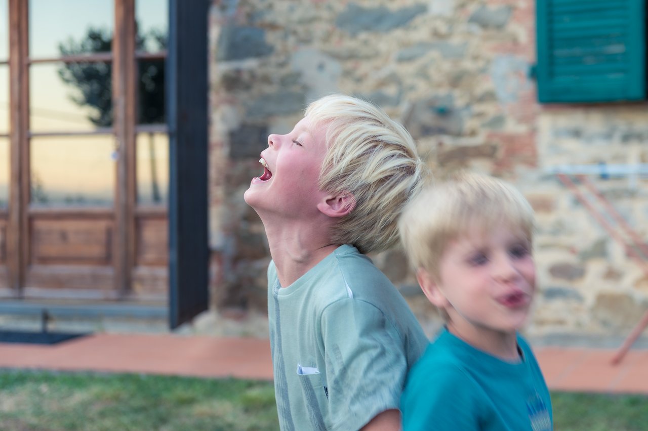 Two children with blonde hair laughing and playing outside near a stone building.