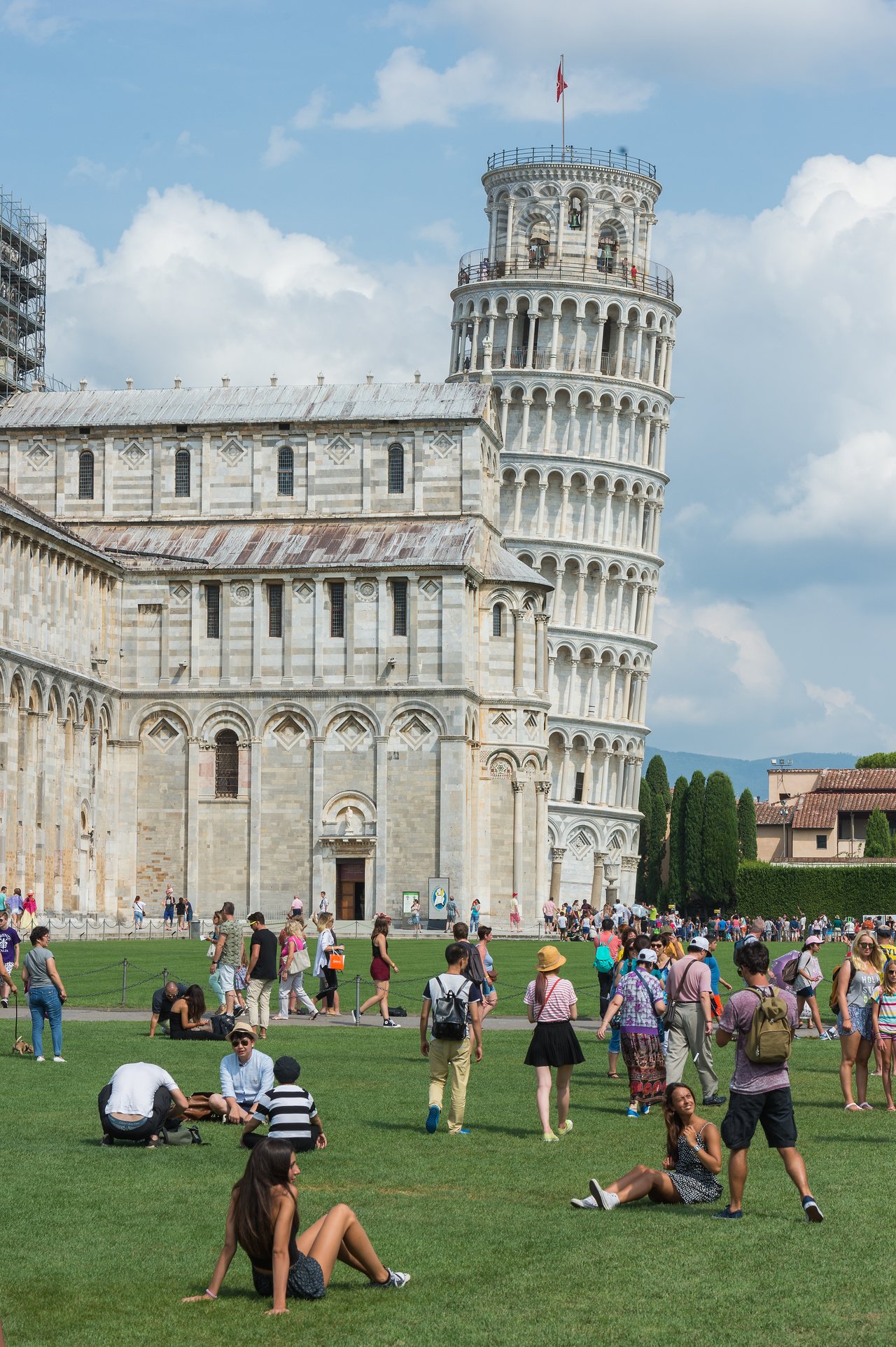 The Leaning Tower of Pisa stands beside a historic building, with many tourists walking and relaxing on the grass.