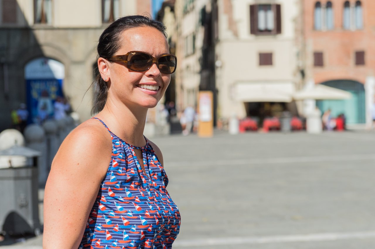 A woman wearing sunglasses and a patterned dress smiles while walking in a sunny public square in Lucca.