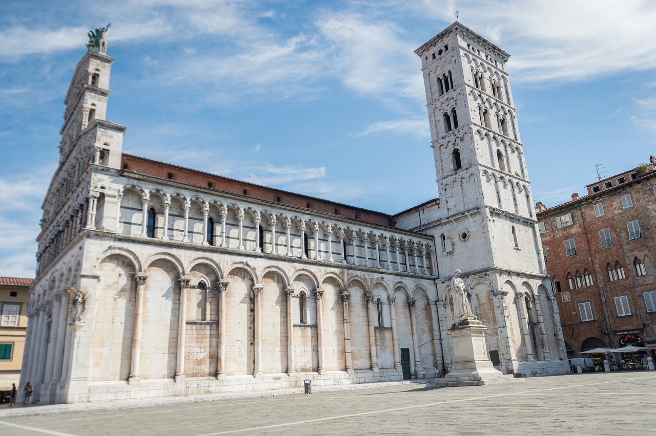 A historic church with a tall bell tower and arched facade in Lucca, Italy, under a blue sky.