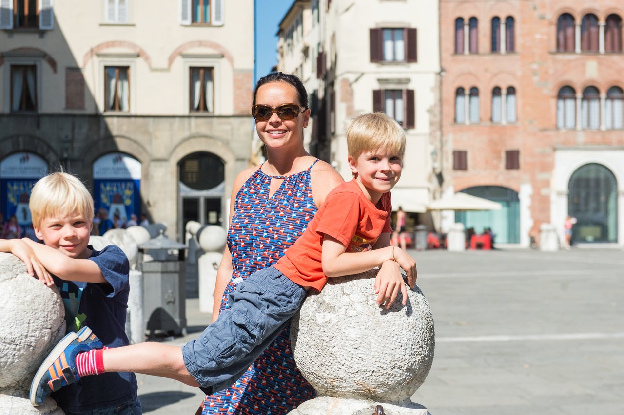 A woman in sunglasses stands smiling while two young boys play on stone bollards in a sunny public square.