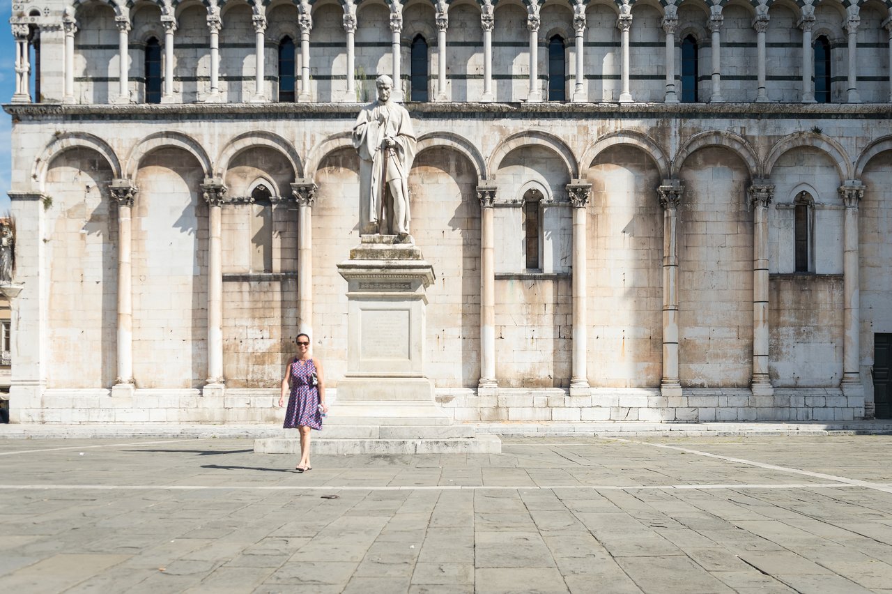 A woman in a purple dress walks past a statue in front of a historic building in Lucca.