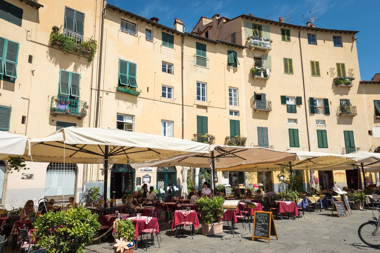 Outdoor café with tables under large umbrellas in a sunny square, surrounded by historic buildings with green shutters.
