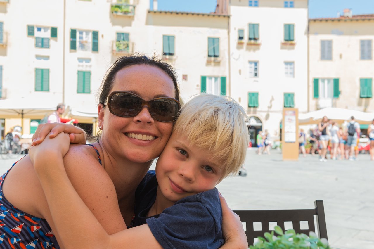 A woman and a young boy hug and smile at an outdoor café in Piazza dell'Anfiteatro, Lucca.