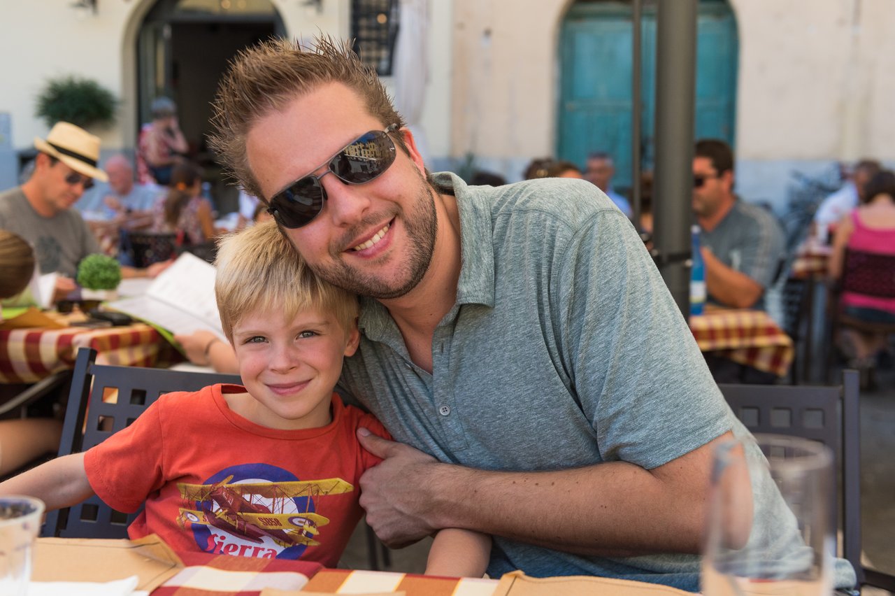 A man and a young boy smile while sitting together at an outdoor café with checkered tablecloths.