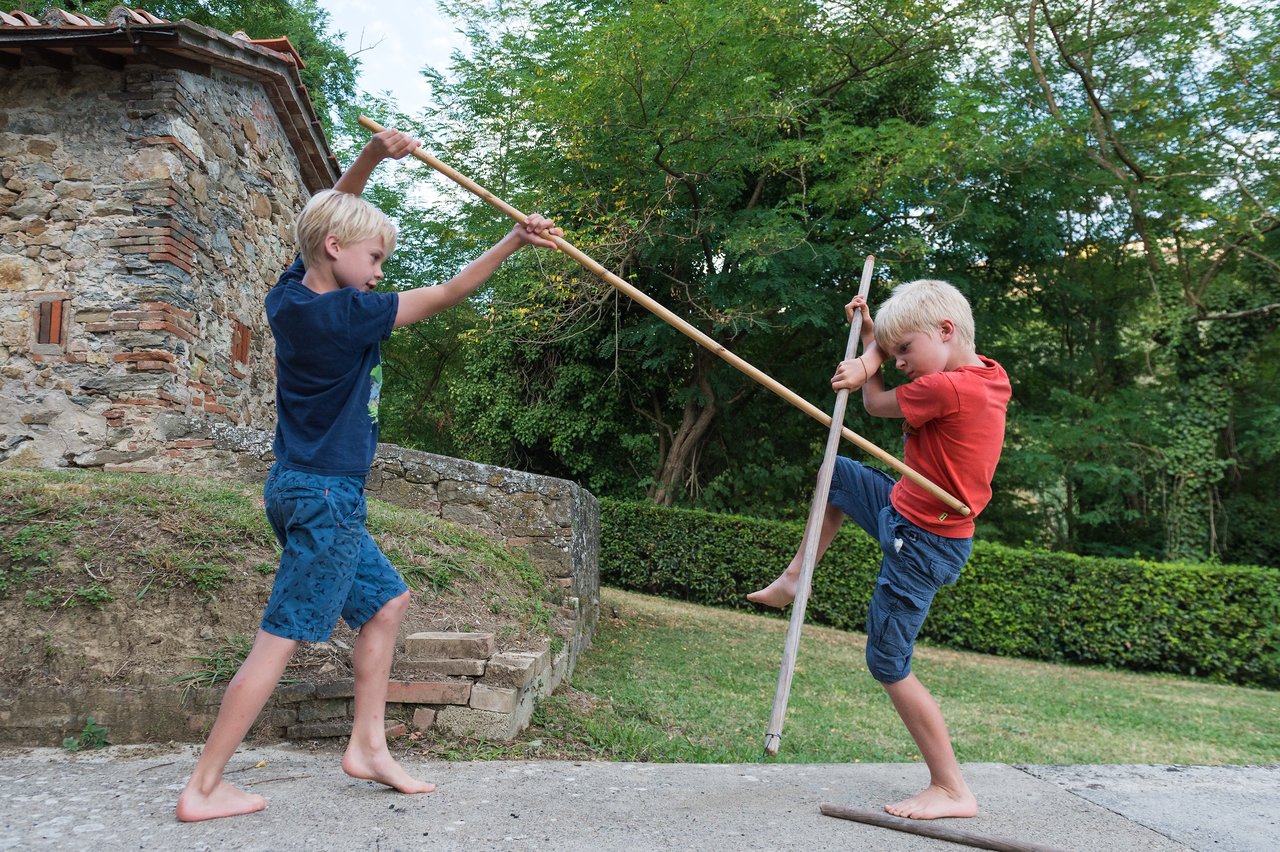 Two barefoot children playfully duel with bamboo sticks outdoors, balancing and striking while smiling.