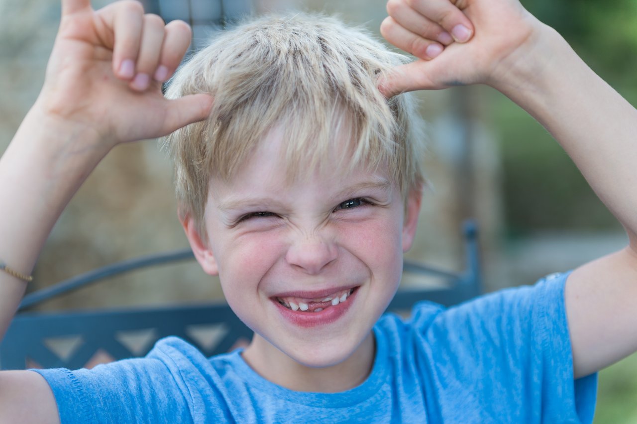 A smiling child in a blue shirt makes a playful gesture with both hands, showing missing front teeth.