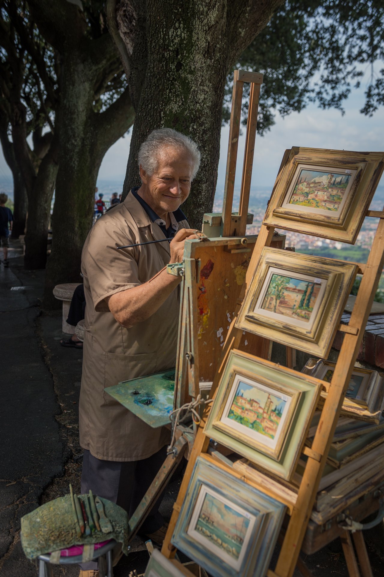 An artist paints outdoors, surrounded by framed landscape paintings displayed on a wooden easel.