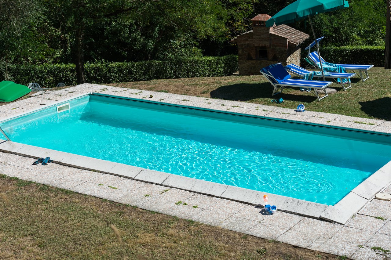 A rectangular swimming pool with clear water, surrounded by lounge chairs, an umbrella, and scattered personal items.