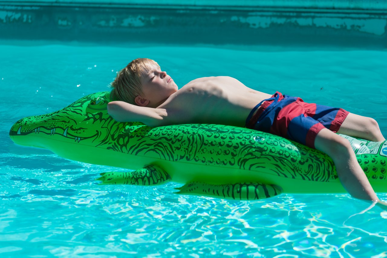 A child in red and blue swim trunks relaxes on a green inflatable alligator in a swimming pool.