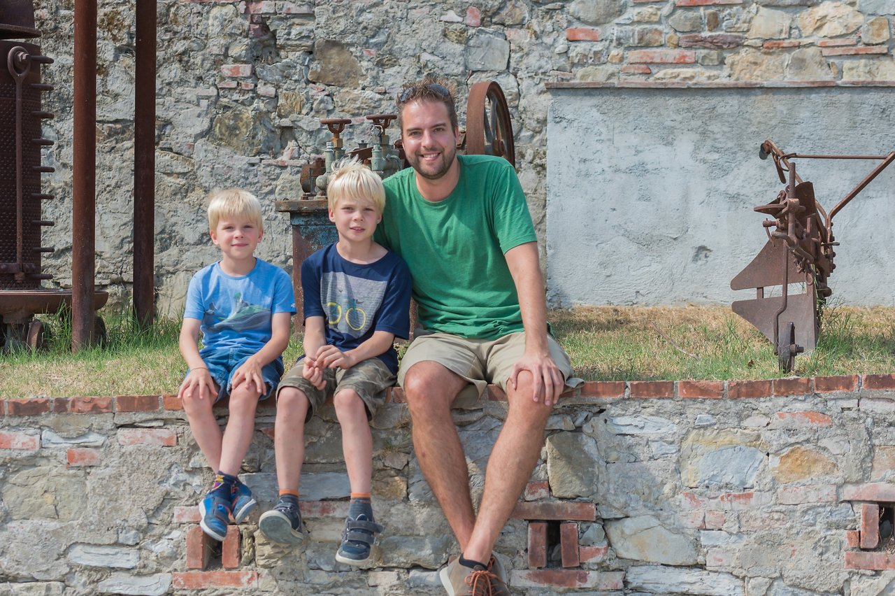 A man and two young boys sit on a stone ledge, smiling at the camera in an outdoor setting.