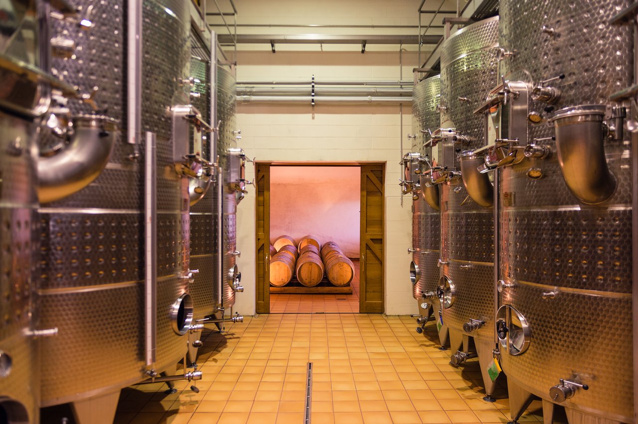 Large metal wine fermentation tanks line a room, leading to a doorway with wooden barrels in the background.