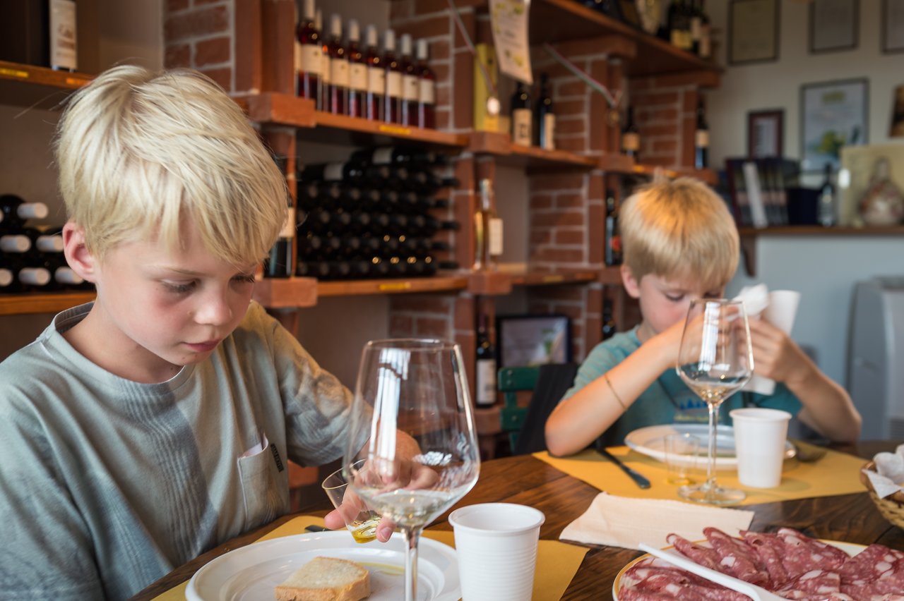 Two children sit at a table with food and wine glasses in a winery, eating and focusing on their meals.
