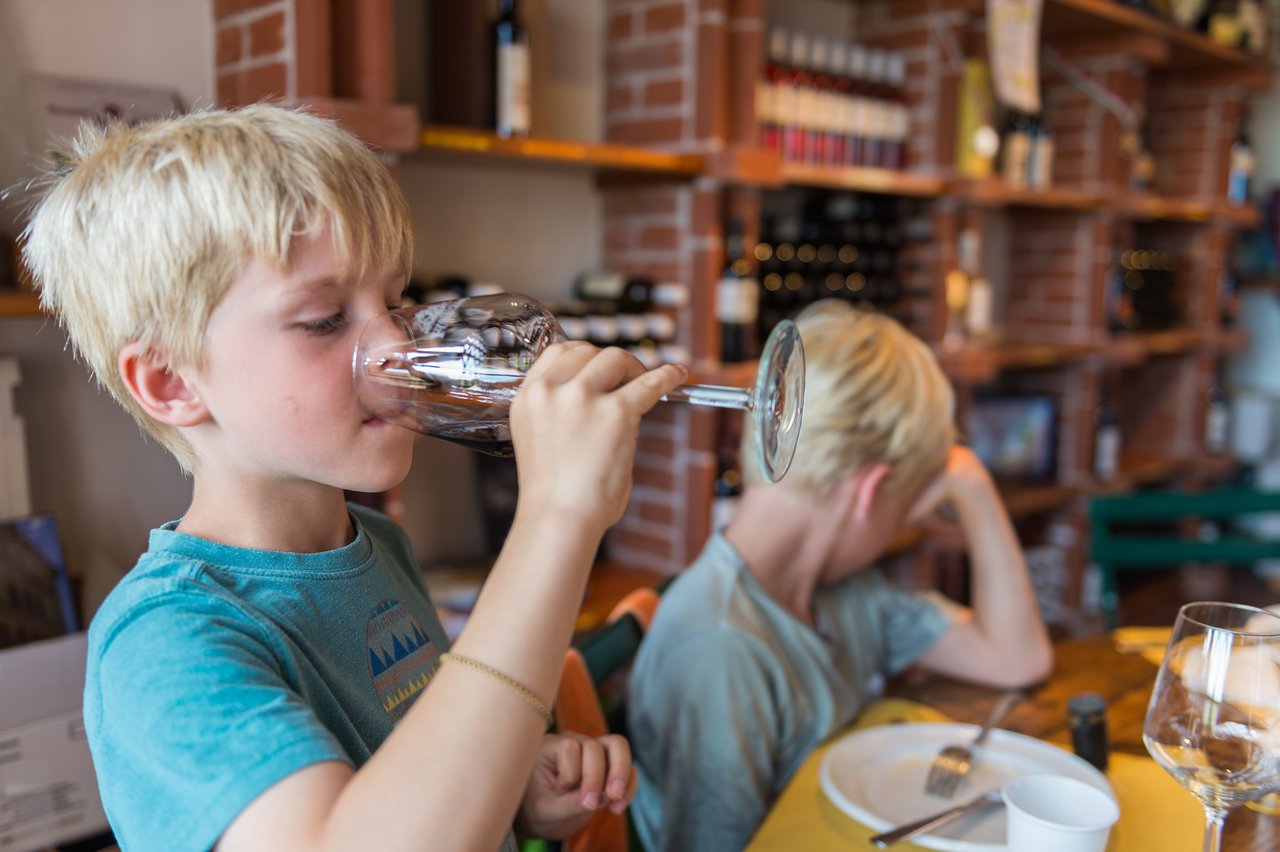 A young child drinks from a wine glass in a tasting room with wine bottles on shelves in the background.