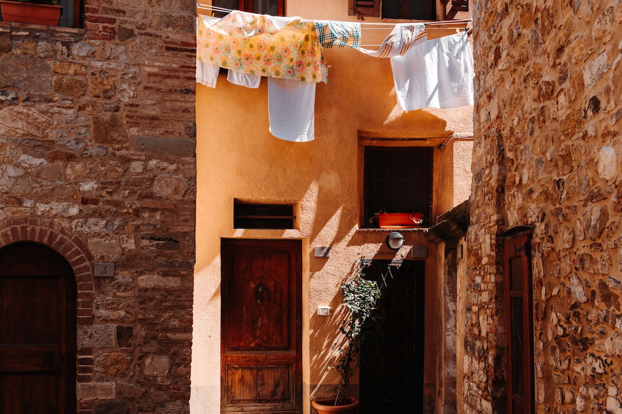 Clothes and linens hang on a clothesline above a doorway in a narrow alley with stone and stucco walls.