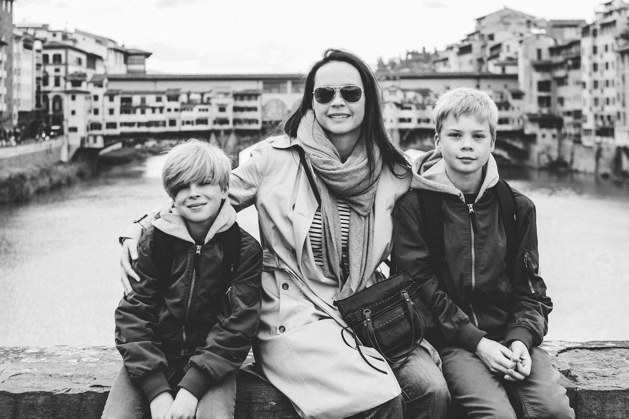 A woman sits between two boys, smiling at the camera with her arms around them, with the Ponte Vecchio behind them.