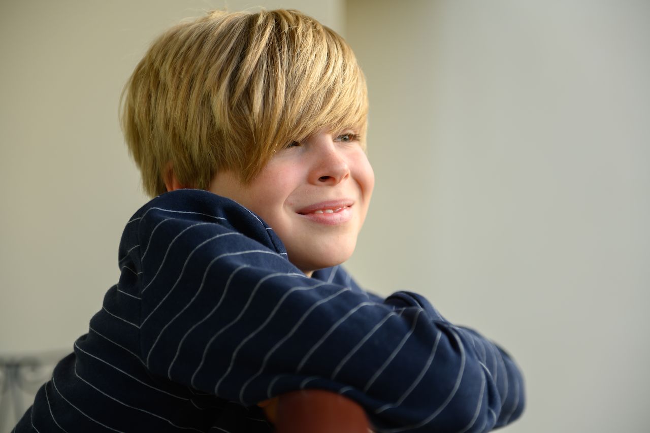 A smiling child with blond hair leans on a chair, looking off to the side.