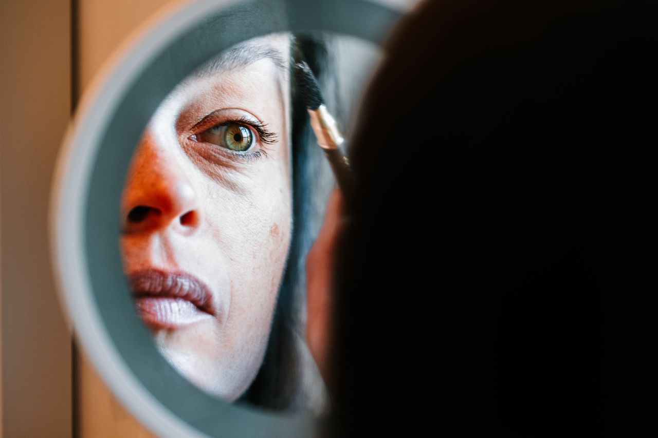 A woman applies make-up to her eyebrow while looking into a round mirror.
