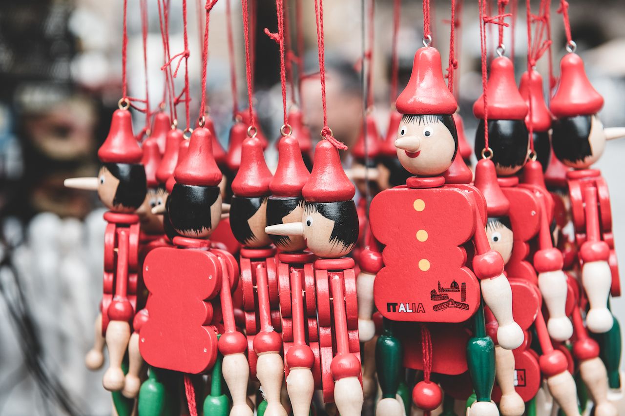 A group of wooden Pinocchio puppets with red hats and strings hang closely together in a market display.