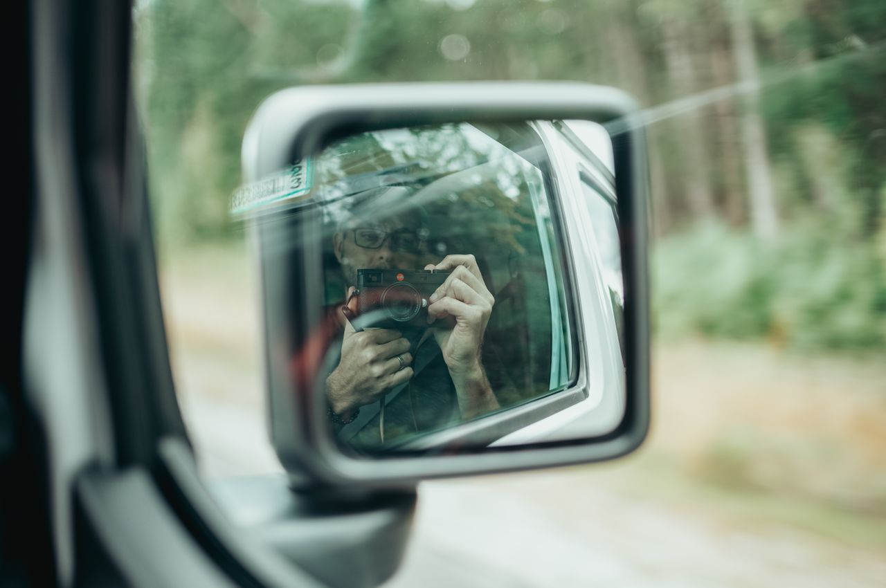 A person takes a selfie with a camera, reflected in the side mirror of a Jeep.