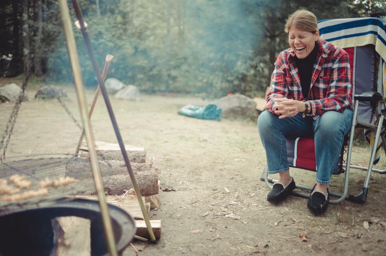 A woman in a plaid shirt sits in a camping chair, laughing with her eyes closed near a fire pit.