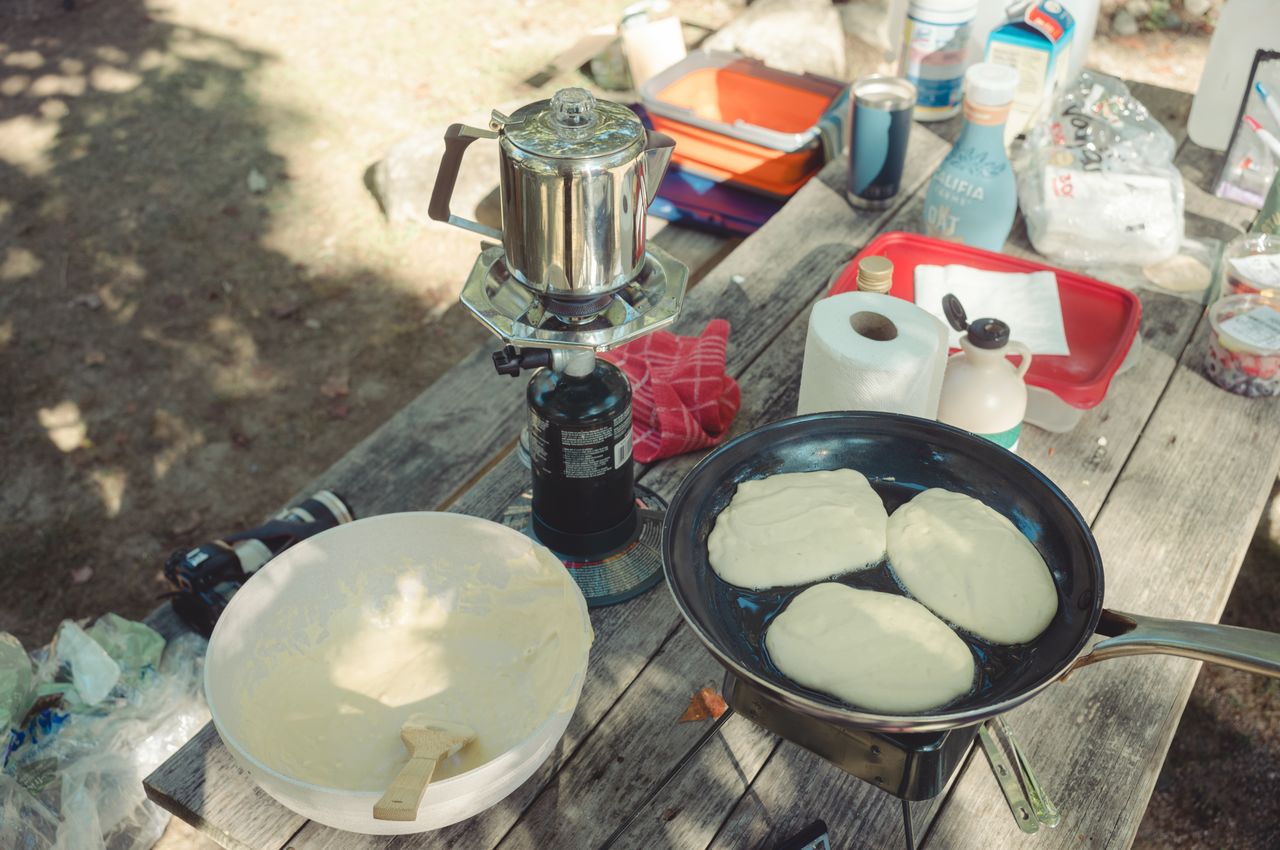 A frying pan on a camping stove with pancakes cooking, surrounded by pancake batter, syrup, and other breakfast items.