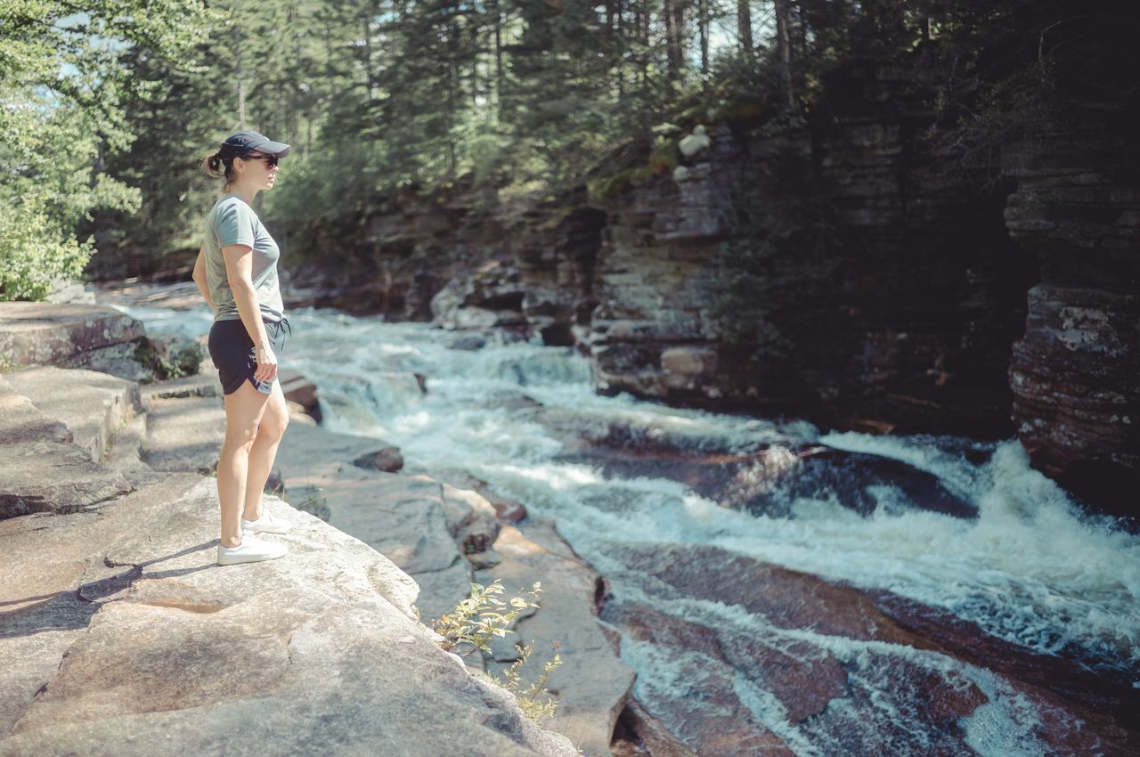 A person stands on a rocky edge, looking at rushing whitewater rapids in a forested area.