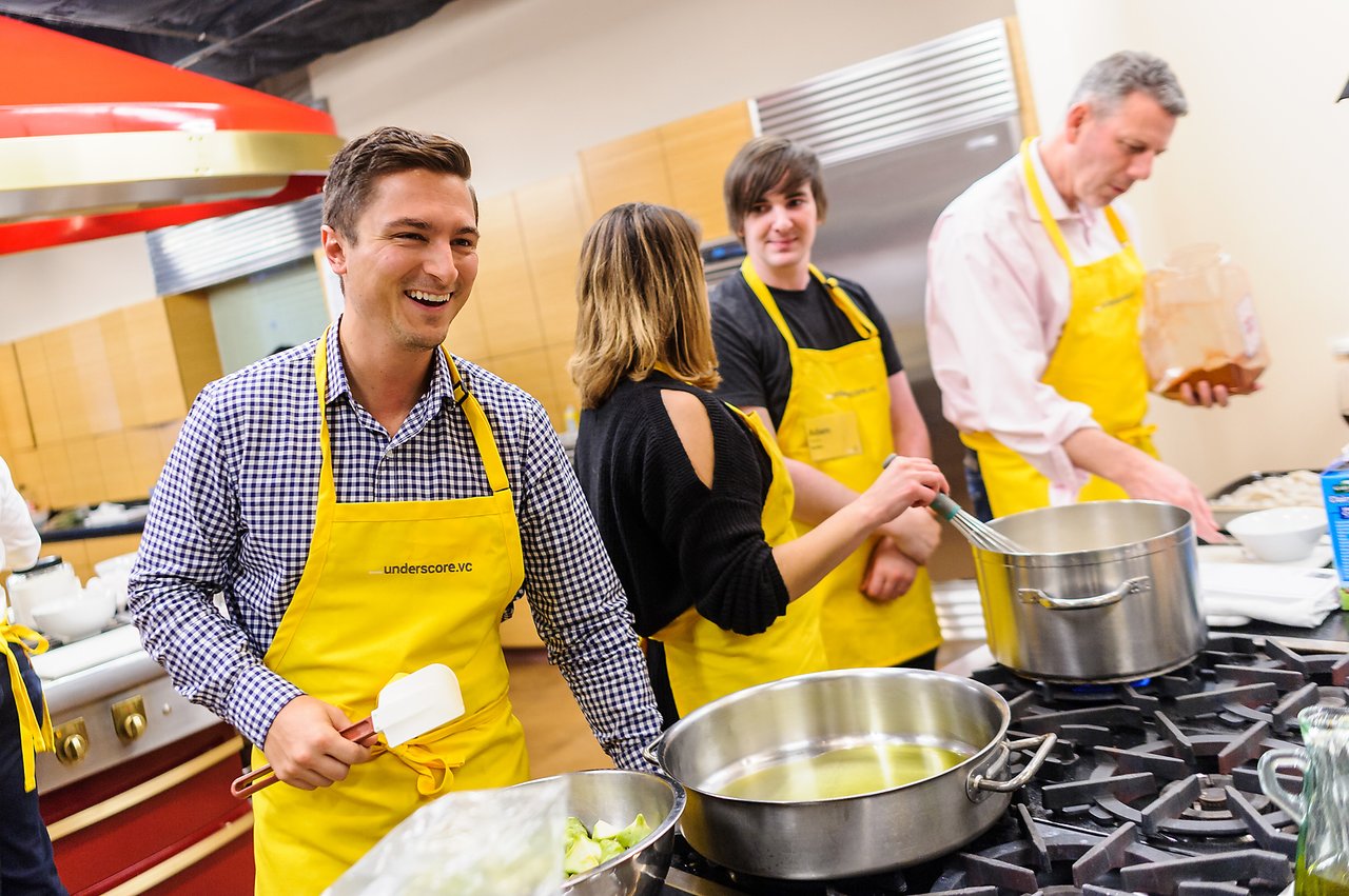 A group of people wearing yellow aprons cook together in a kitchen, stirring pots and preparing ingredients.