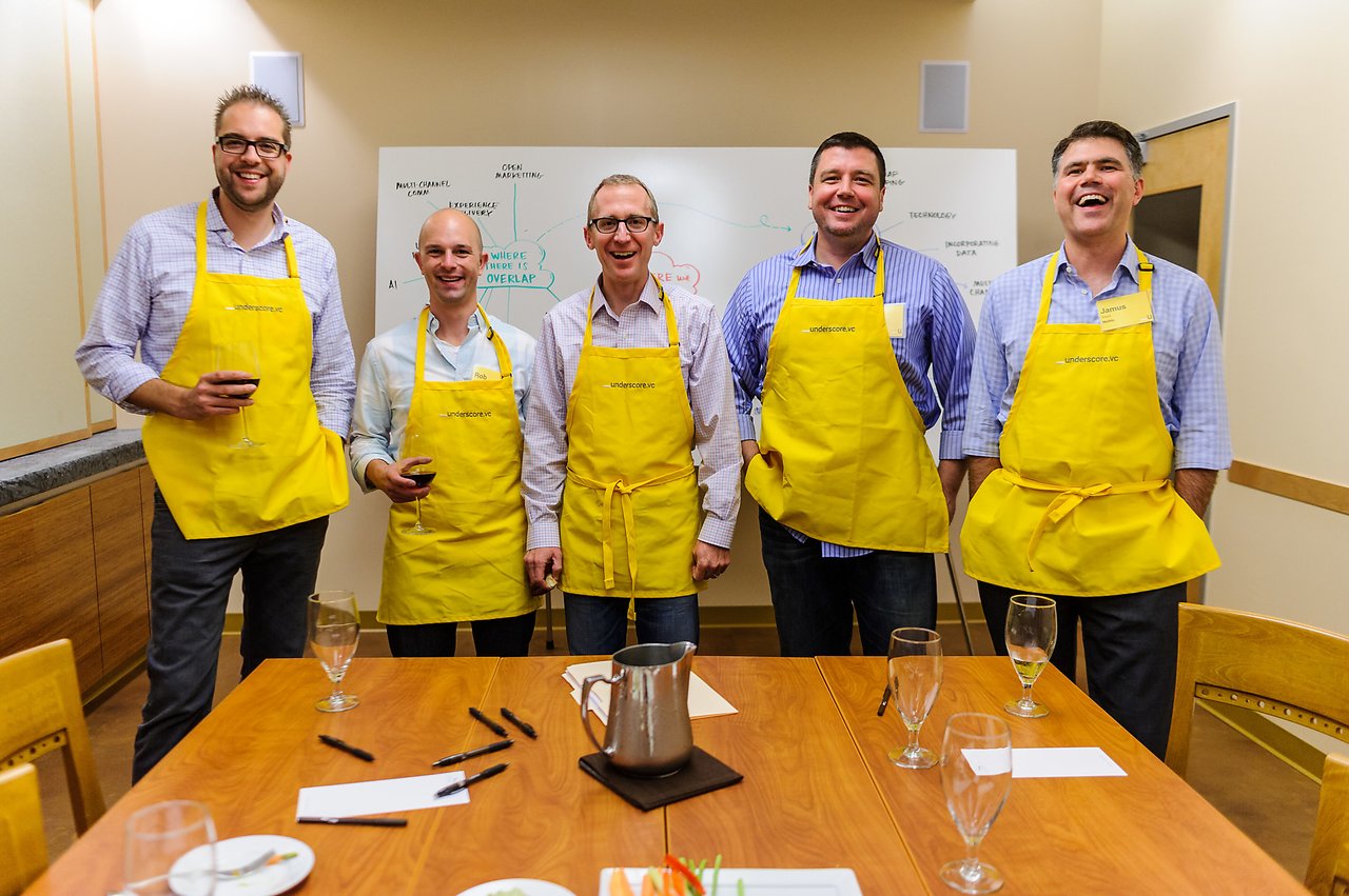 Five people wearing yellow aprons stand together, smiling and holding drinks in a workshop setting.
