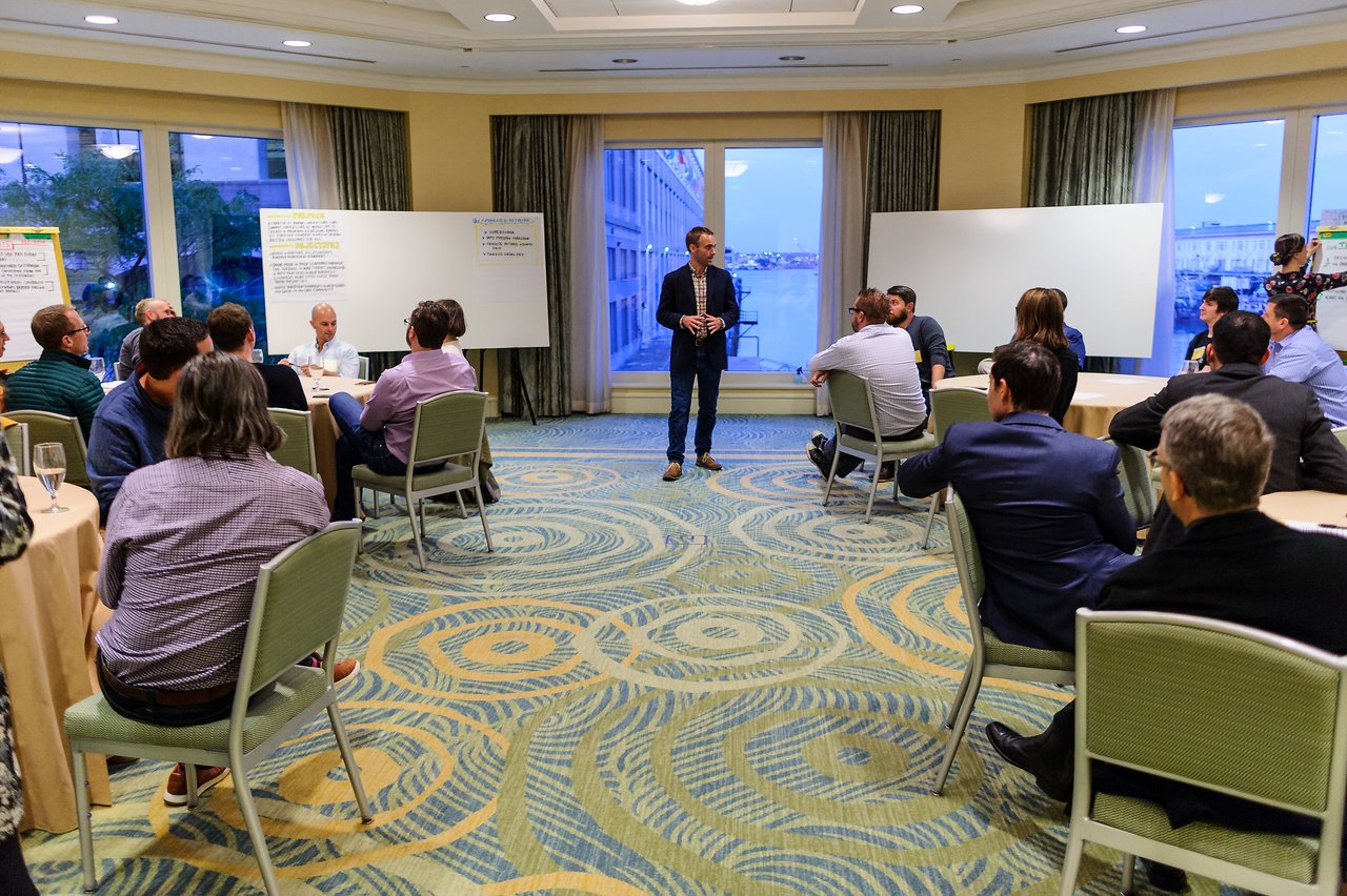 A man stands in the center of a conference room, speaking to seated participants during a workshop discussion.