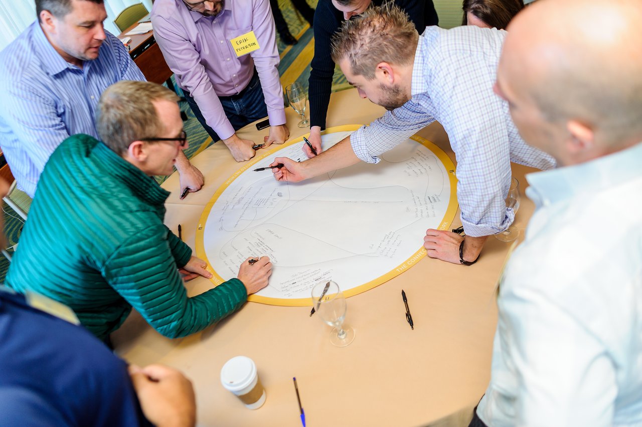 A group of people collaborates around a table, writing and discussing ideas on a large circular paper.