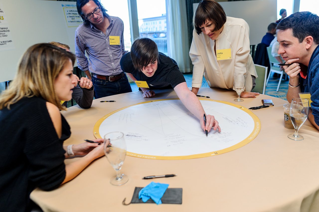 A group of people collaborates around a table, with one person writing on a large circular sheet.