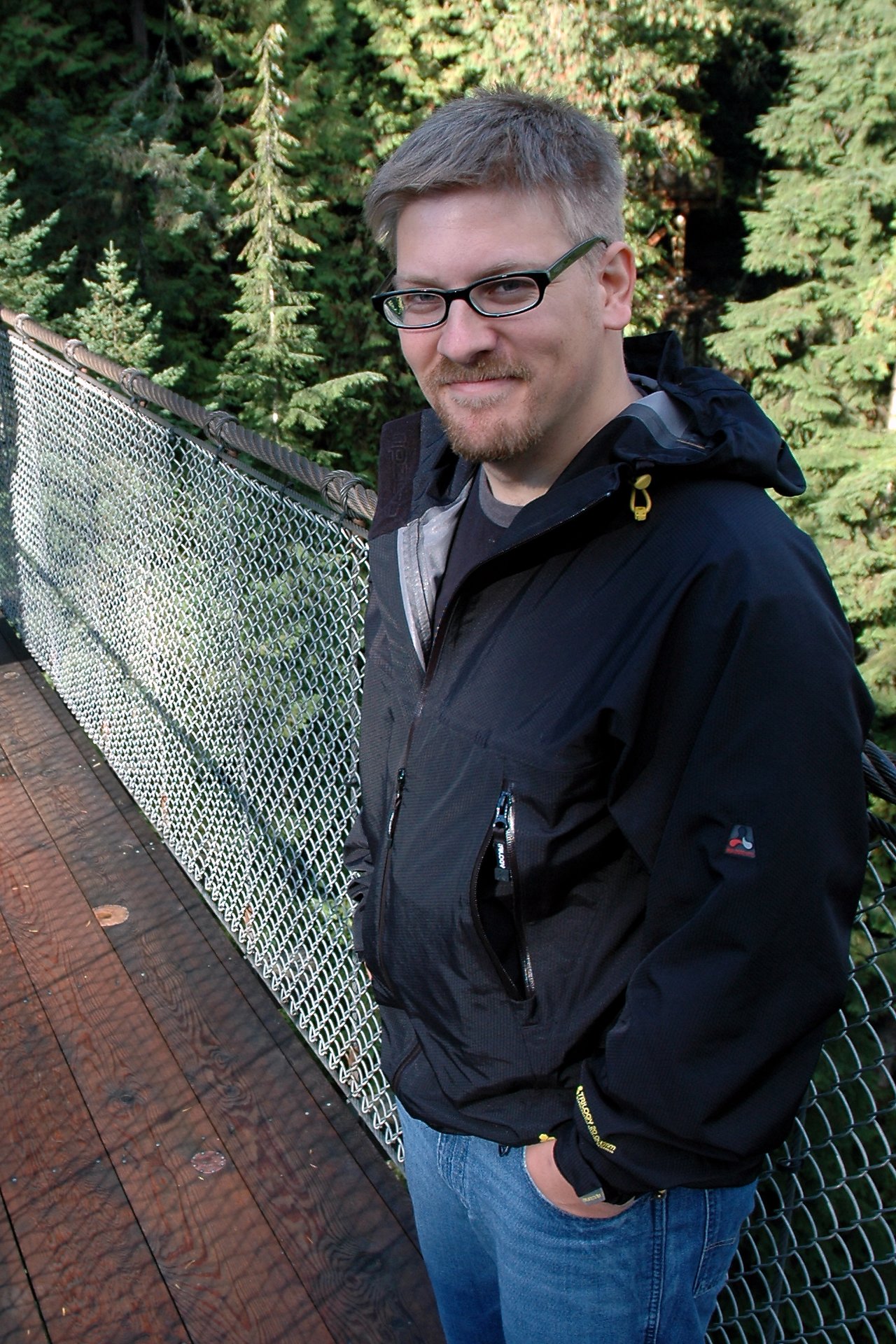 A man wearing glasses and a black jacket stands on a suspension bridge, smiling at the camera.