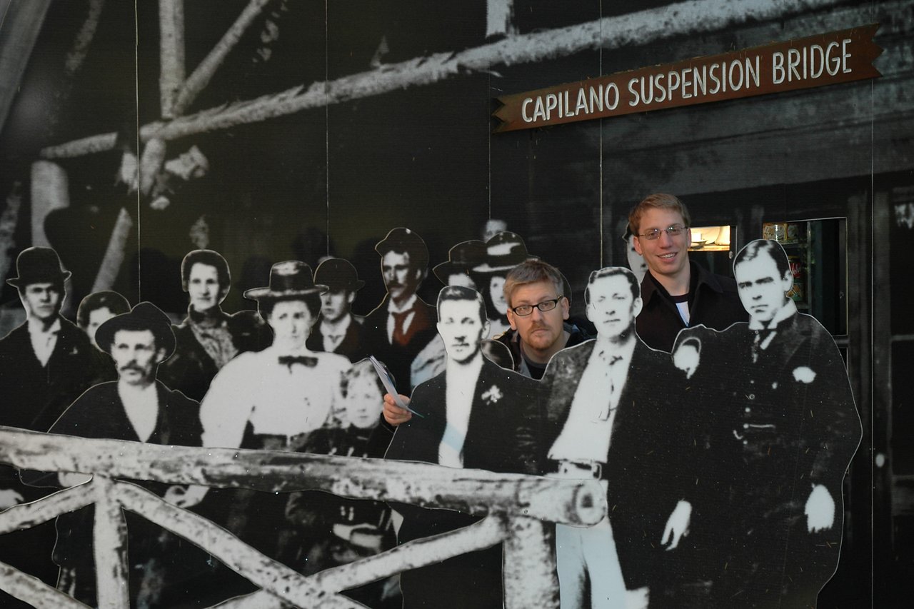 Two people pose behind a historical cutout display at Capilano Suspension Bridge, blending into the black-and-white scene.