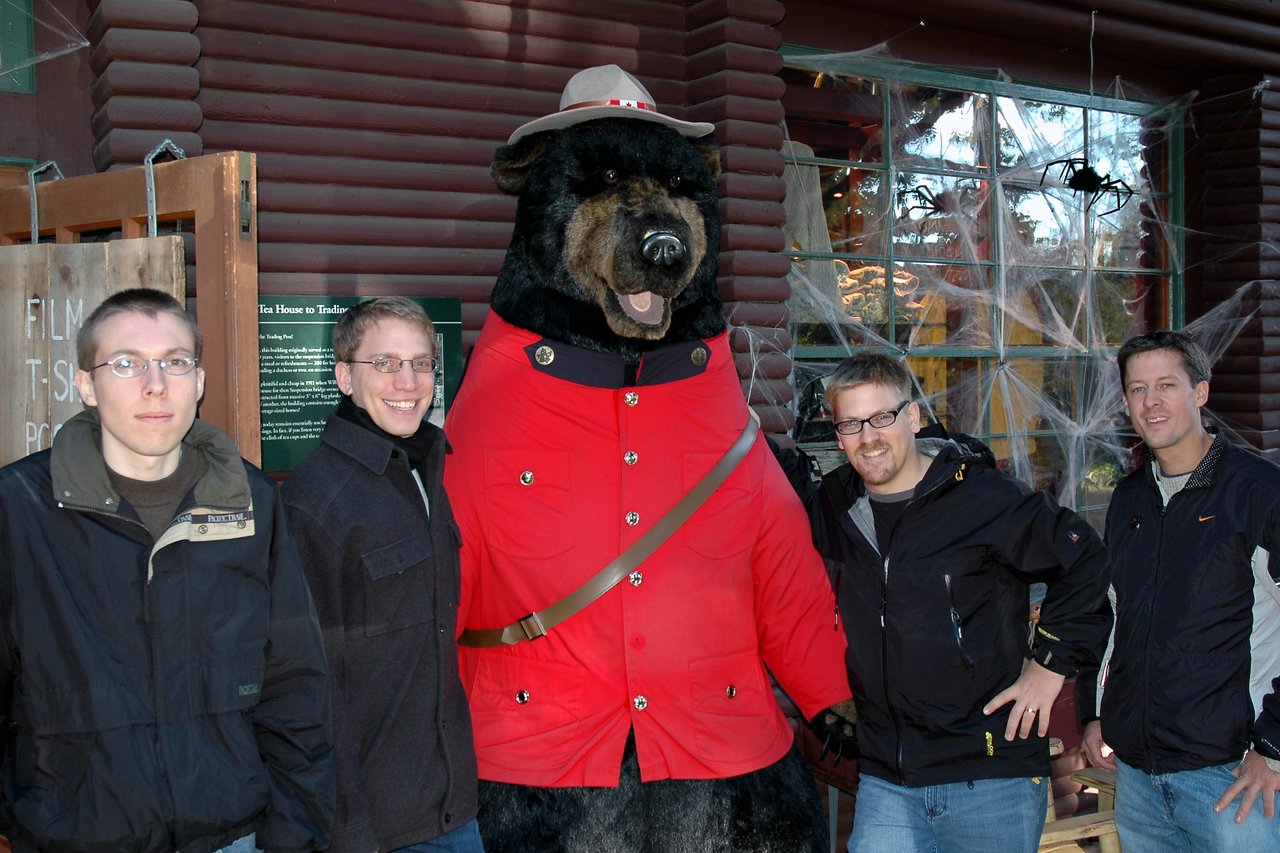 Four men stand next to a large bear statue dressed as a Mountie, posing for a group photo outdoors.