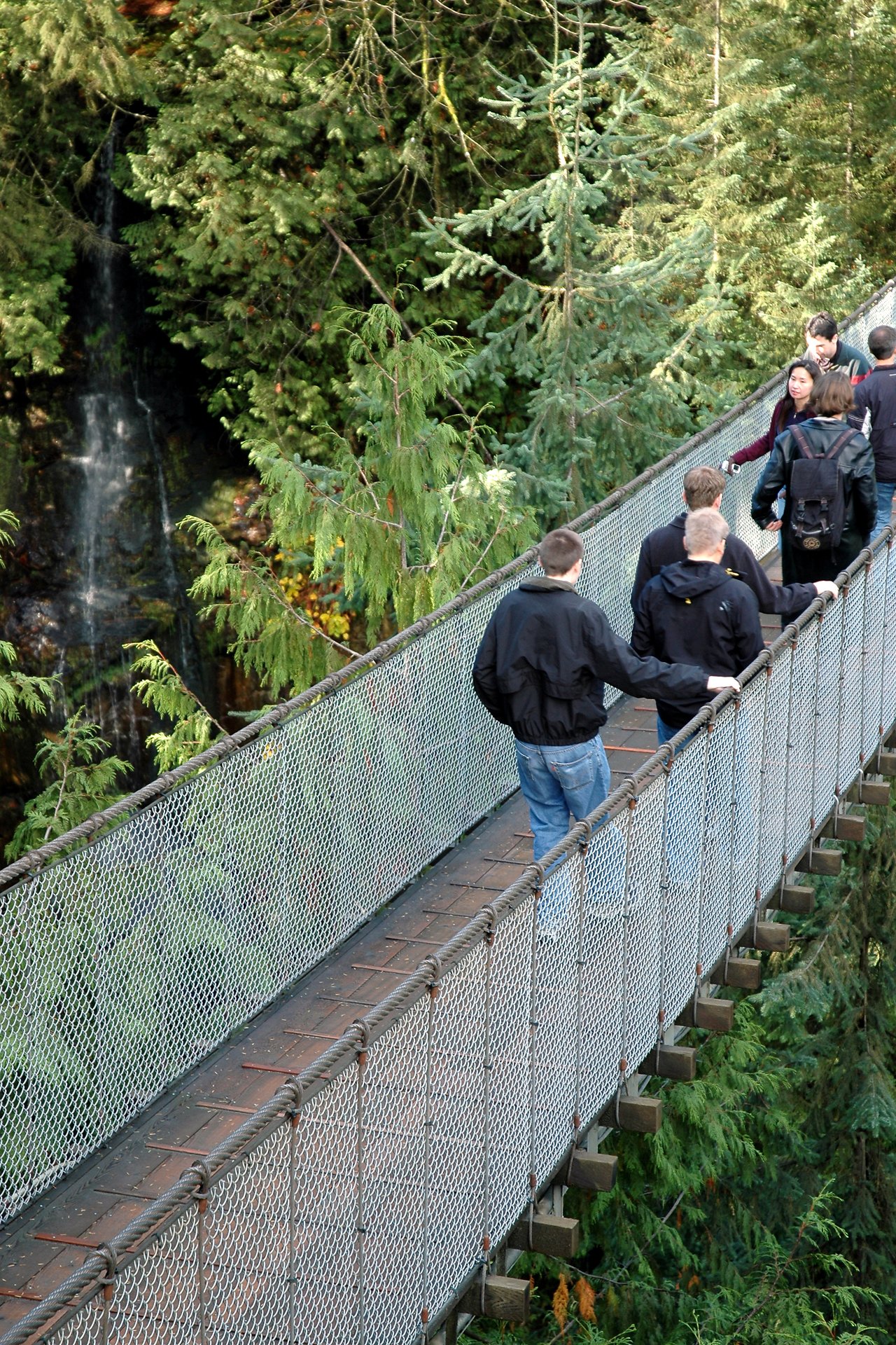 People walk across the Capilano Suspension Bridge, surrounded by tall trees and a waterfall in the background.