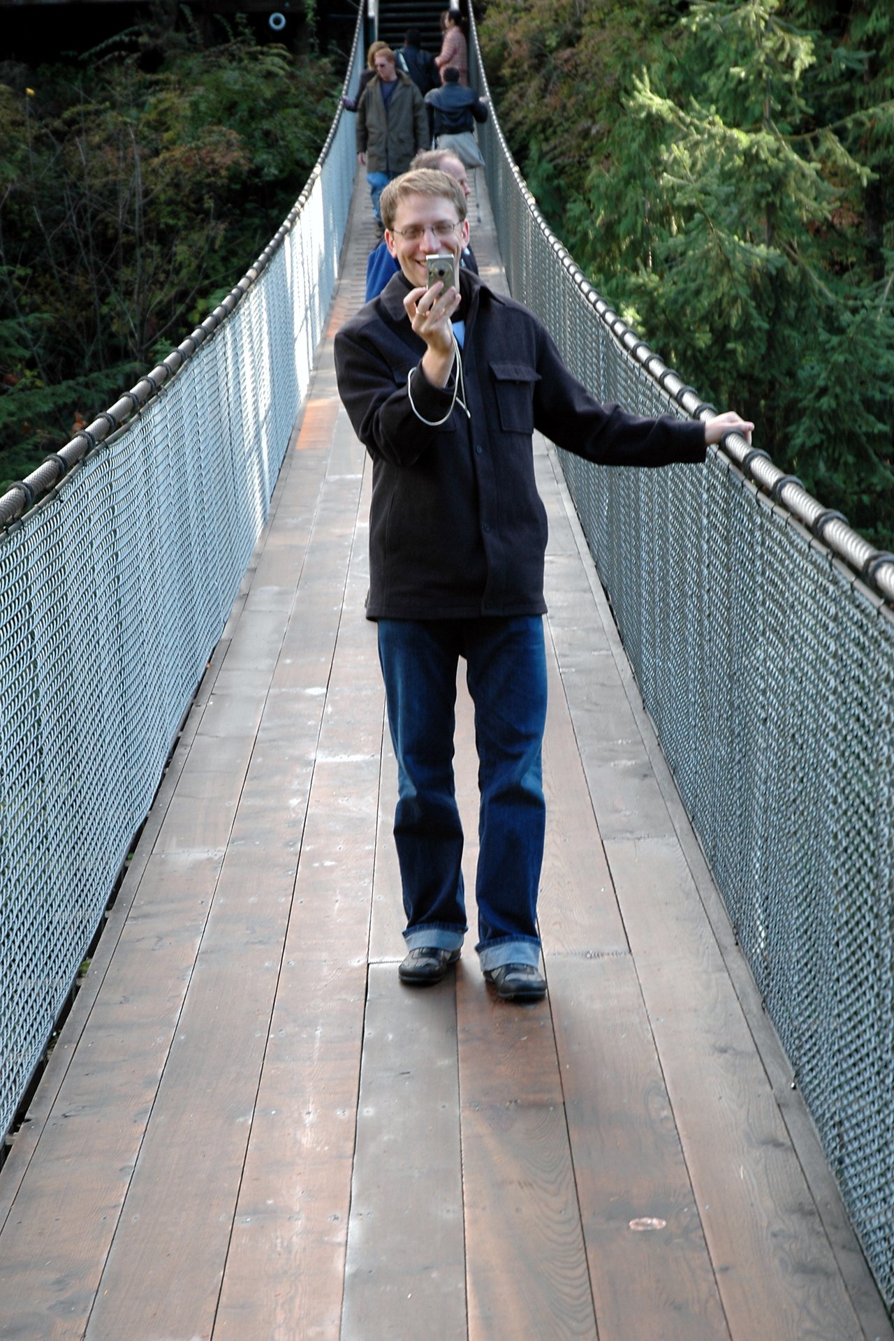 A man stands on a suspension bridge, smiling while taking a photo with a small camera.