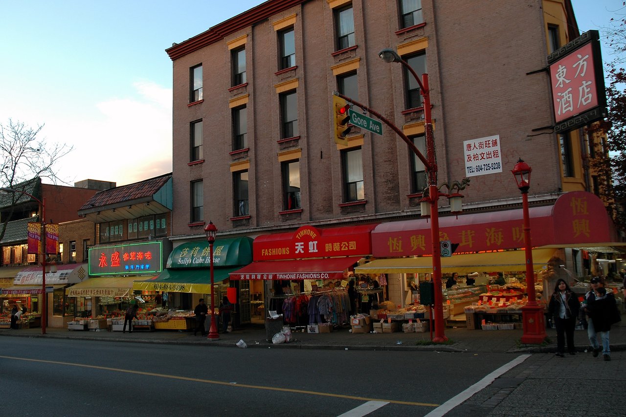 Street view of Chinatown with shops, market stalls, and pedestrians walking past colorful storefronts under a traffic light.