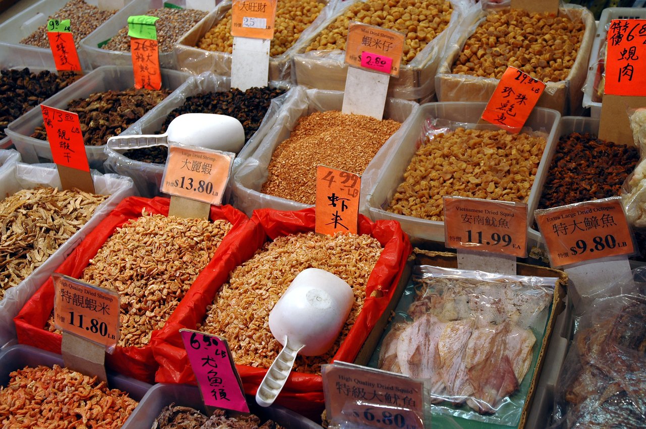 Bins of dried seafood with price tags and a white scoop at a market in Chinatown.