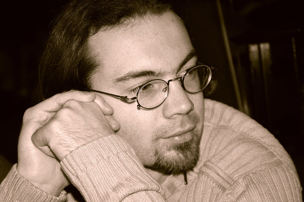 A man wearing glasses and a sweater rests his head on his hands, looking off to the side.