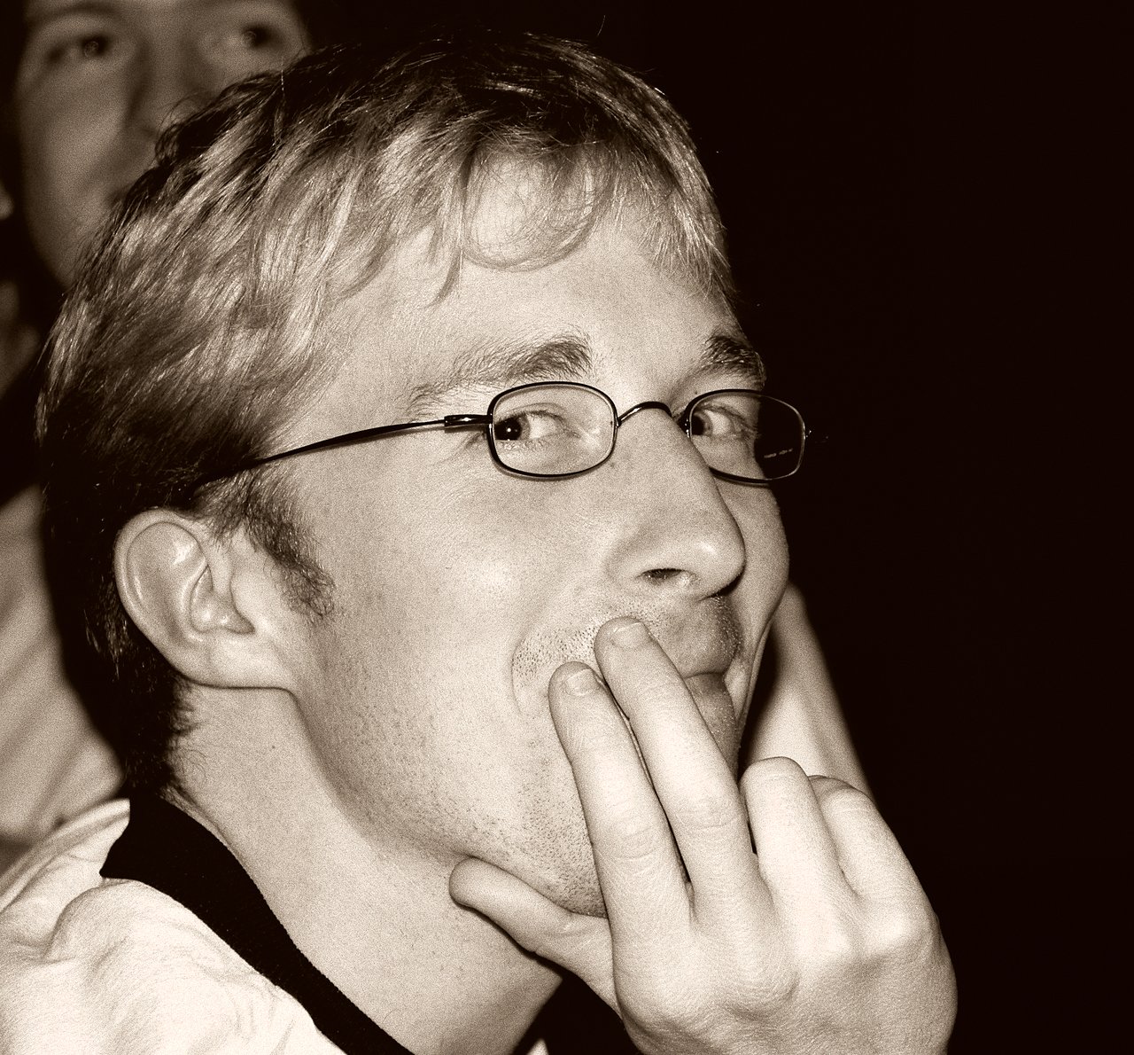 A person wearing glasses smiles playfully while resting their fingers on their lips at a social gathering.
